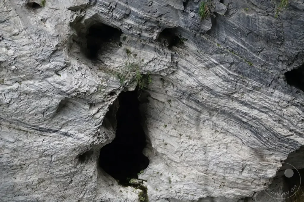 Im Taroko Nationalpark in Taiwan offenbart sich die beeindruckende Formation von Felsen mit sichtbaren Gesteinsschichten. Eine dunkle Höhle ragt aus der Felswand hervor und verleiht der Szenerie eine geheimnisvolle Atmosphäre. Die raue Schönheit der Natur ist in den Strukturen des Gesteins und dem Spiel von Licht und Schatten eingefangen.