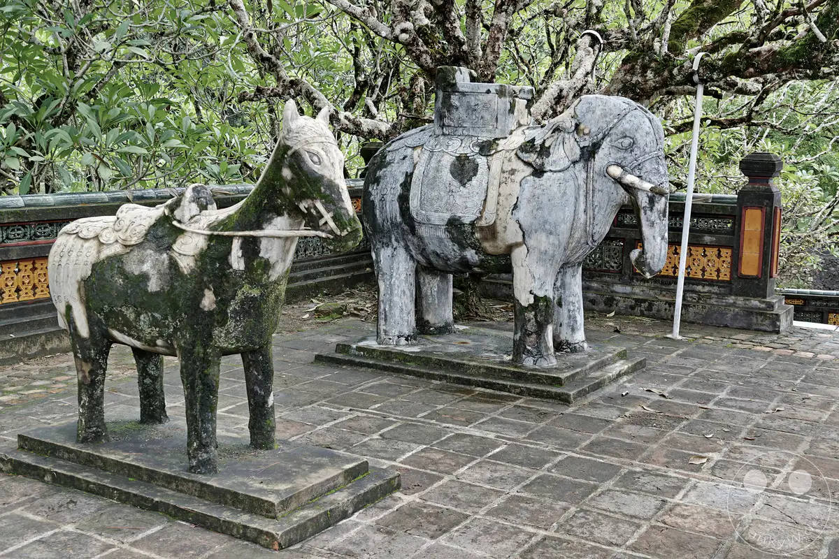Ein steinernes Elefanten- und Pferdestatuenpaar, das von Moos und Flechten überzogen ist, steht auf einem gepflasterten Hof in Hue, Vietnam. Die Statuen befinden sich vor einer kunstvoll verzierten Mauer und unter dem dichten Blätterdach alter Bäume, was eine ehrwürdige und zugleich leicht melancholische Atmosphäre erzeugt. Die kunstvoll gestalteten steinernen Tiere sind ein markantes Merkmal der weitläufigen Anlage der kaiserlichen Gräber.