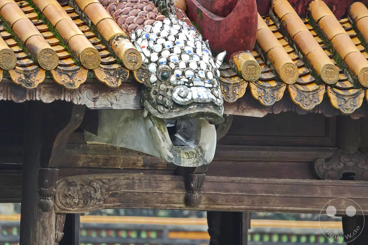 Eine detaillierte Ansicht der ornamentalen Dachkonstruktion des Grabmals von Tu Duc in Hue, Vietnam. Die aufwendige Verzierung mit keramischen Elementen und die traditionellen gelben Dachziegel verleihen der Szene eine historische und kulturelle Tiefe. Moosbewuchs auf den Ziegeln deutet auf das Alter und die Wetterbeständigkeit der Architektur hin.