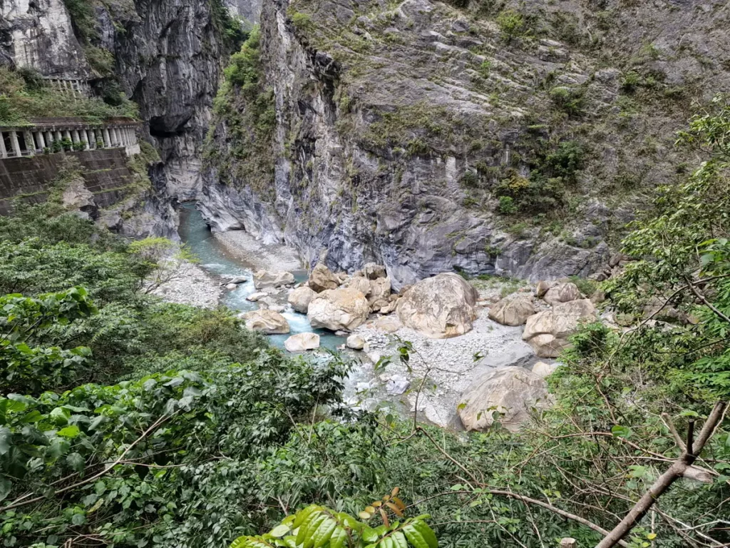 Der Liwu River schlängelt sich durch die tiefe Taroko-Schlucht in Taiwan, umgeben von steilen, zerklüfteten Felswänden. Große Felsbrocken säumen das Flussbett, während üppige grüne Vegetation die unteren Hänge und den Vordergrund des Bildes bedeckt. Die Szenerie vermittelt ein Gefühl von Erhabenheit und der unberührten Kraft der Natur.
