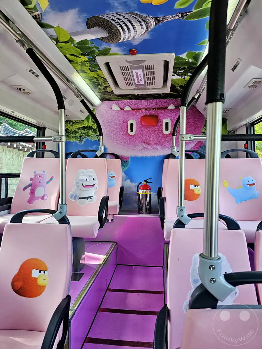 Ein pinkfarbener Bus mit Cartoon-Motiven und Sitzbezügen ist in Seoul, Südkorea zu sehen. Die bunte Innenausstattung mit Charakteren und einer Nachbildung des Namsan-Turms schafft eine spielerische Atmosphäre, die besonders für Kinder ansprechend ist. Die lila Beleuchtung und die Dekorationen verleihen dem Innenraum des Busses eine fröhliche und einladende Stimmung.