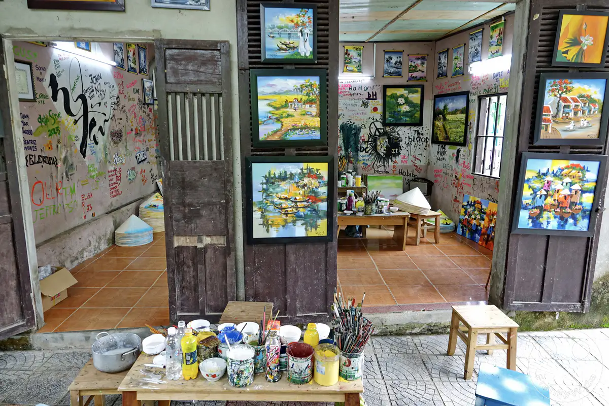 Eine belebte Kunstwerkstatt in Hue, Vietnam, präsentiert eine Vielzahl von handbemalten Bildern und Kunsthandwerk auf den Wänden und Tischen. Die Szenerie strahlt eine kreative und handwerkliche Atmosphäre aus, die zum Entdecken lokaler Kunst und Souvenirs einlädt. Farbenfrohe Werke und künstlerische Utensilien schaffen ein lebendiges Bild der vietnamesischen Kunstszene.