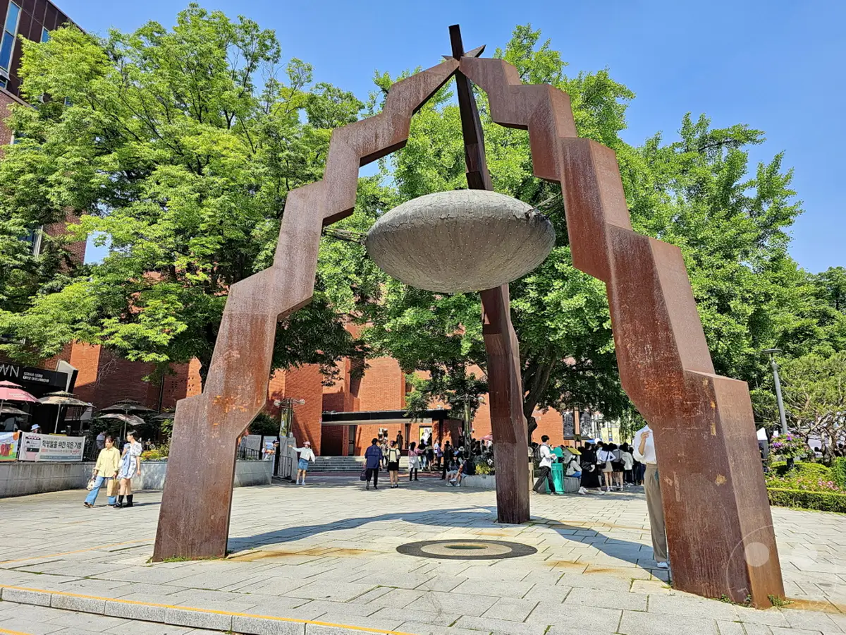 Eine markante, rostbraune Metallskulptur dominiert den Marronnier Park im Daehangno district von Seoul, Südkorea. Umgeben von üppigem Grün und an einem sonnigen Tag gelegen, lädt das Kunstwerk zum Verweilen ein. Menschen bewegen sich entspannt im Hintergrund und tragen zur lebendigen Atmosphäre des städtischen Parks bei.