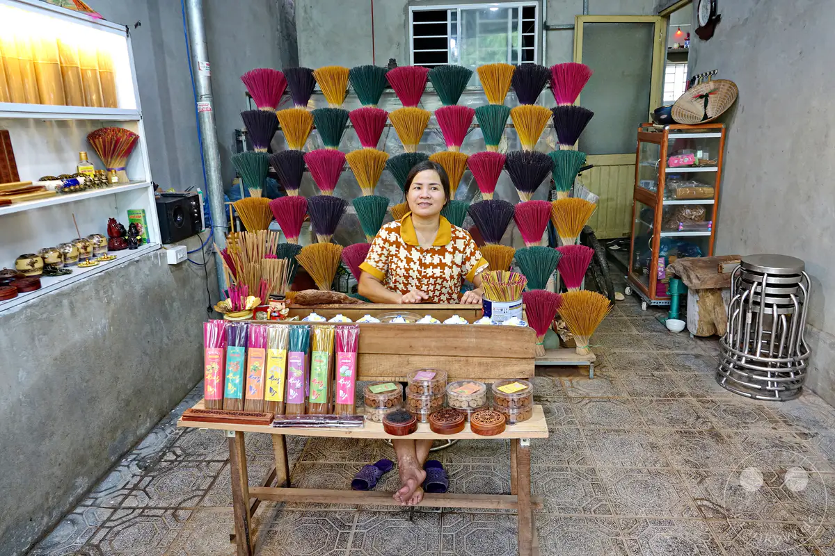Eine Frau sitzt hinter einem Tisch, der mit bunten Räucherstäbchen und verschiedenen Behältern gefüllt ist, in ihrem Geschäft in Hue, Vietnam. Überall sind sorgfältig gestapelte Bündel von Räucherstäbchen in verschiedenen Farben ausgestellt, die eine lebendige und traditionelle Atmosphäre schaffen. Die Szene strahlt die Ruhe und Hingabe der Handwerkskunst aus, die für die Herstellung von Räucherstäbchen typisch ist.