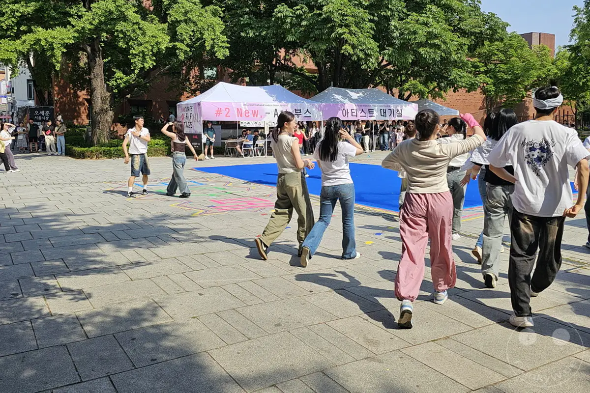 Menschen versammeln sich in Südkorea im Daehangno district, genauer gesagt im Marronnier Park, um an einem modernen Kunstfestival teilzunehmen. Die Atmosphäre ist lebhaft, während Besucher die verschiedenen Ausstellungen und Darbietungen erkunden. Mehrere Zelte und farbenfrohe Bodengestaltungen deuten auf ein dynamisches kulturelles Ereignis im Herzen von Seoul hin.