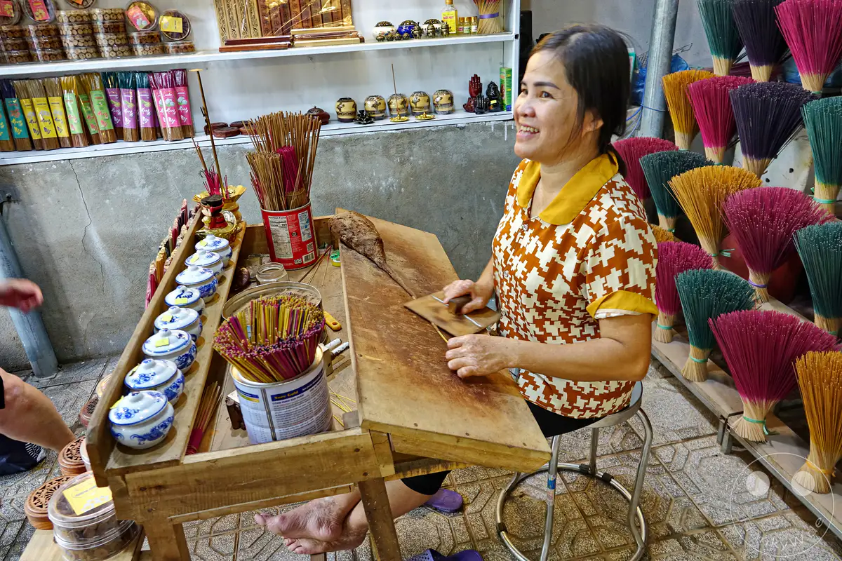 Eine Frau in Vietnam, in der Stadt Hue, stellt traditionell Räucherstäbchen her. Die bunten Räucherstäbchen und die geschäftige Atmosphäre des Handwerks vermitteln eine lebendige und authentische Stimmung. Verschiedene Arten von Räucherstäbchen und dazugehörige Behälter sind zu sehen.