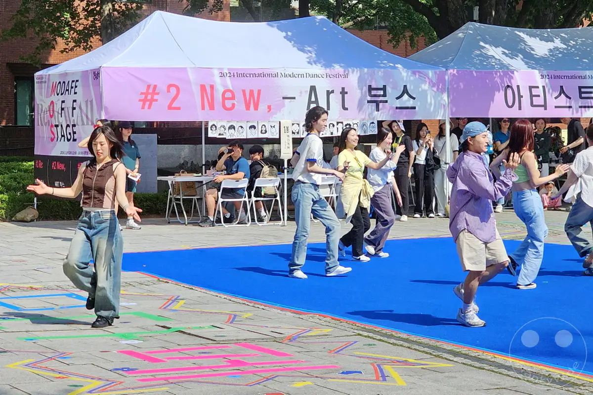 Eine Gruppe von Tänzern, die bei einem modernen Tanzfestival im Marronnier Park im Daehangno district von Seoul, Südkorea, auftritt. Die Darbietung findet unter freiem Himmel unter einem Festzelt statt, das mit der Aufschrift "2024 International Modern Dance Festival" gekennzeichnet ist. Die fröhliche Atmosphäre wird durch die bunten Kostüme der Tänzer und die lebhaften Posen unterstrichen.