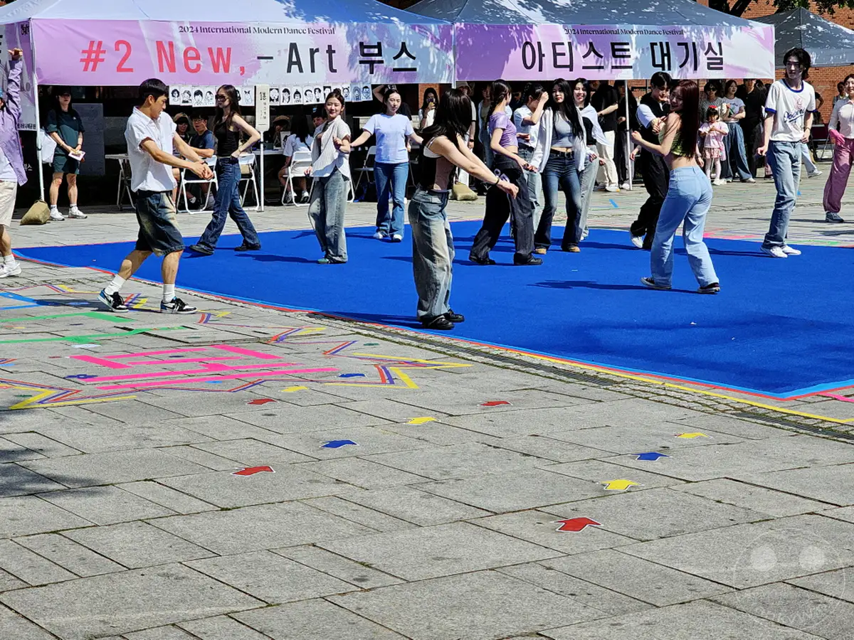 Menschen tanzen auf einem blauen Teppich im Rahmen des "2023 International Modern Dance Festival" in Südkorea. Die Szene spielt sich im Daehangno district von Seoul ab, wahrscheinlich im Marronnier Park, wo bunte Bodenmarkierungen für die Aufführung genutzt werden. Die Atmosphäre ist belebt und künstlerisch, im Freien unter einem Pavillon.