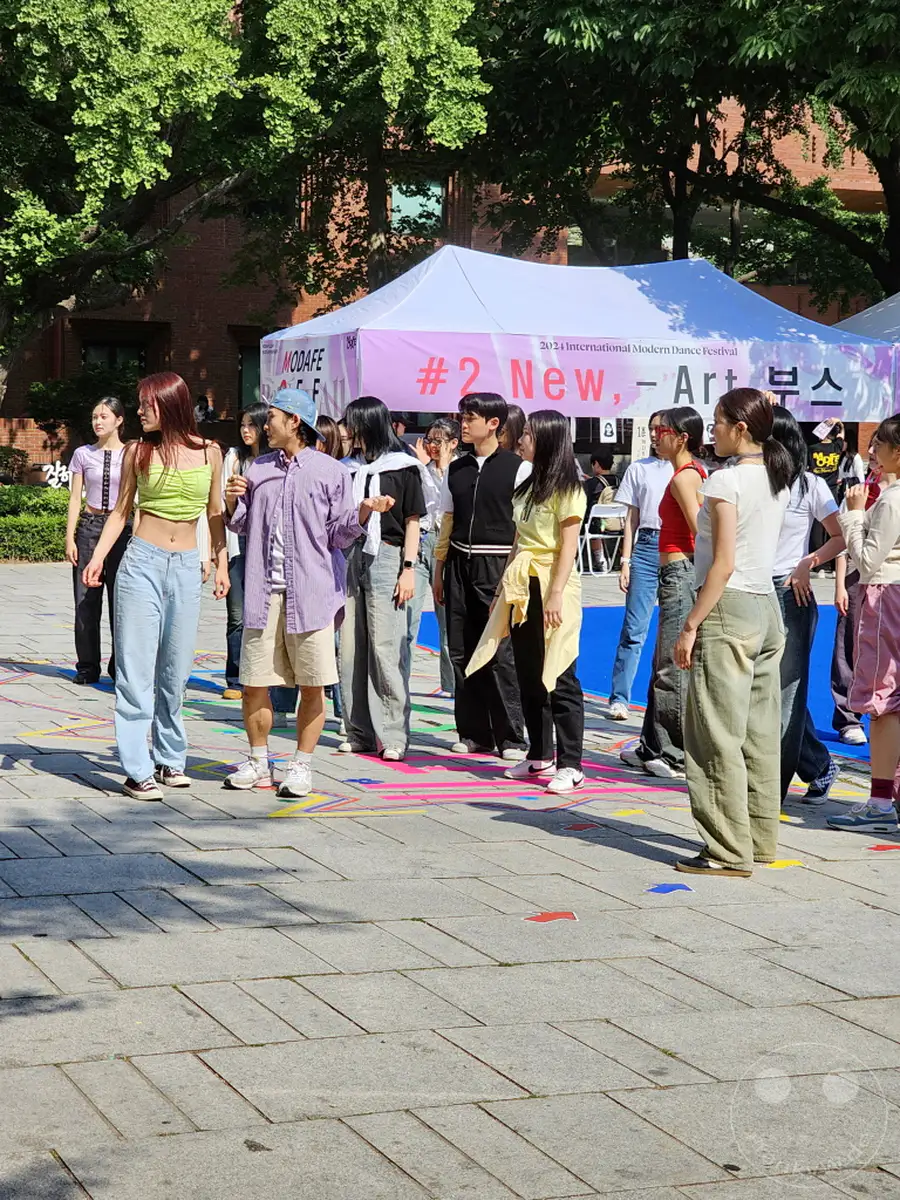 Eine Gruppe junger Menschen versammelt sich auf einem sonnigen Platz in Südkorea, Seoul, im Daehangno district, Marronnier Park. Sie stehen vor einem Zelt mit der Aufschrift „International Modern Dance Festival“, gekleidet in lässiger Freizeitkleidung. Die Atmosphäre wirkt entspannt und jugendlich, während sie auf den Beginn einer Veranstaltung warten.