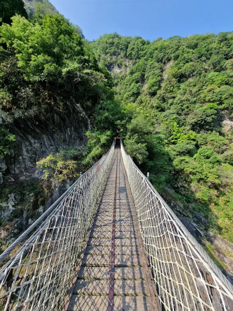Eine robuste Hängebrücke überquert eine tiefe Schlucht im Taroko Nationalpark in Taiwan. Die felsigen, mit üppigem Grün bewachsenen Hänge steigen steil zu beiden Seiten an und rahmen den Weg ein. Der klare blaue Himmel und die klare Luft versprechen eine belebende Wanderung durch diese atemberaubende Naturkulisse.