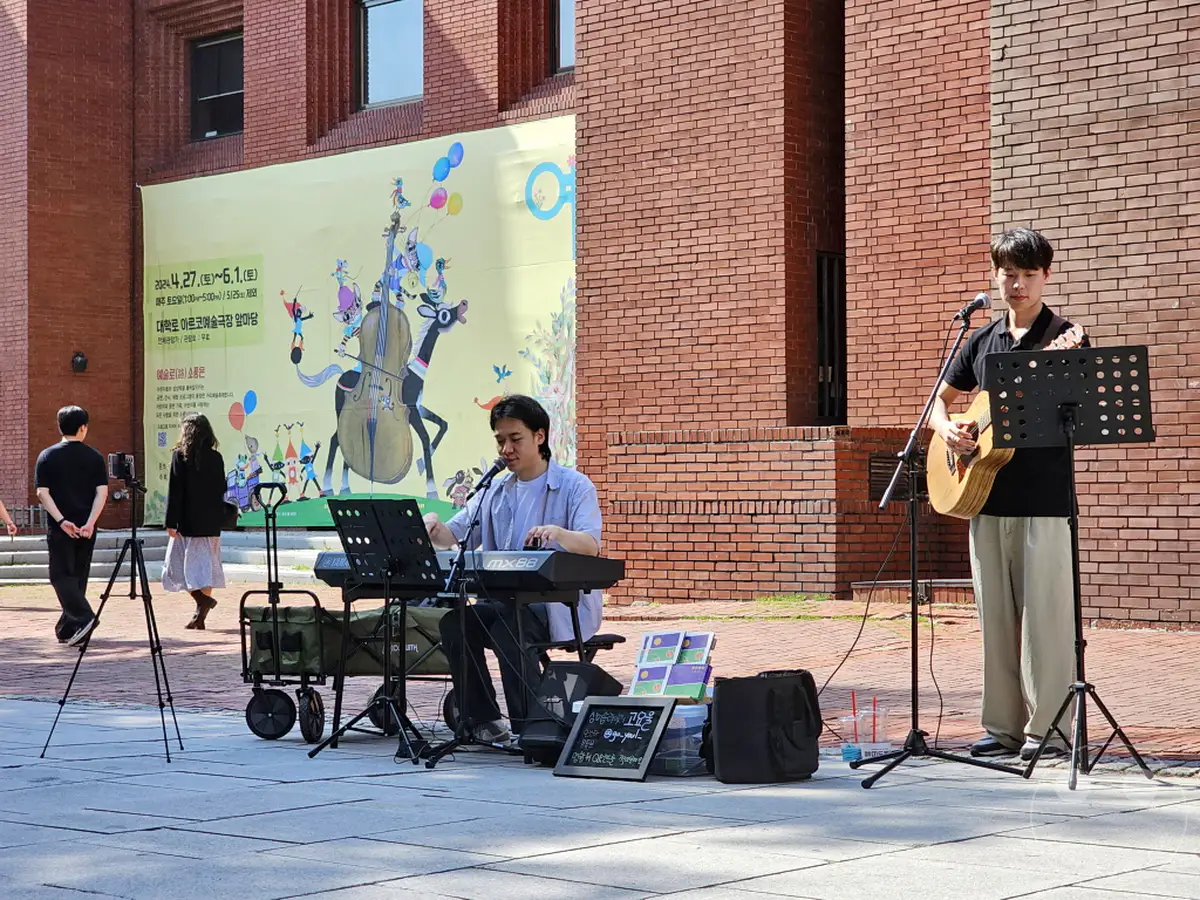 Ein junger Mann spielt Gitarre und singt in Südkorea, Seoul im Daehangno district. Neben ihm sitzt ein anderer Musiker an einem Keyboard. Die Szene strahlt eine entspannte und künstlerische Atmosphäre aus, während die beiden auf der Straße vor einer großen bunten Wandmalerei auftreten.