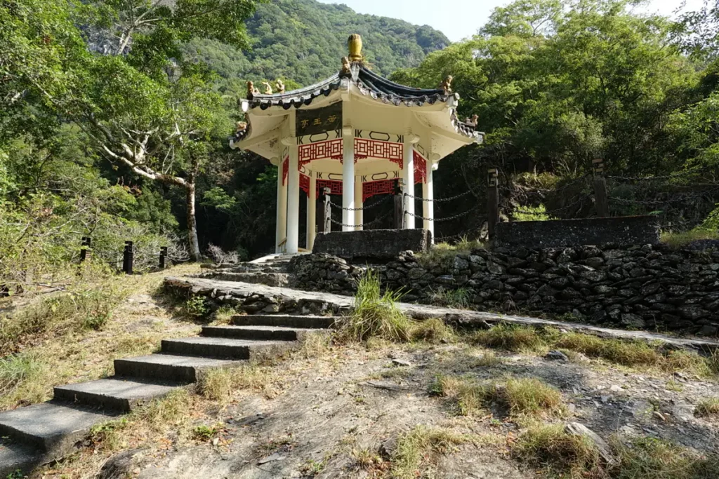 Ein traditioneller chinesischer Pavillon, bekannt als Yuewangting, erhebt sich inmitten der üppigen Vegetation des Taroko Nationalparks in Taiwan. Steintreppen führen zu diesem architektonischen Juwel, das von schattigen Bäumen und einer majestätischen Bergkulisse umgeben ist. Die Szene strahlt eine friedliche und historische Atmosphäre aus, die die natürliche Schönheit der Region widerspiegelt.