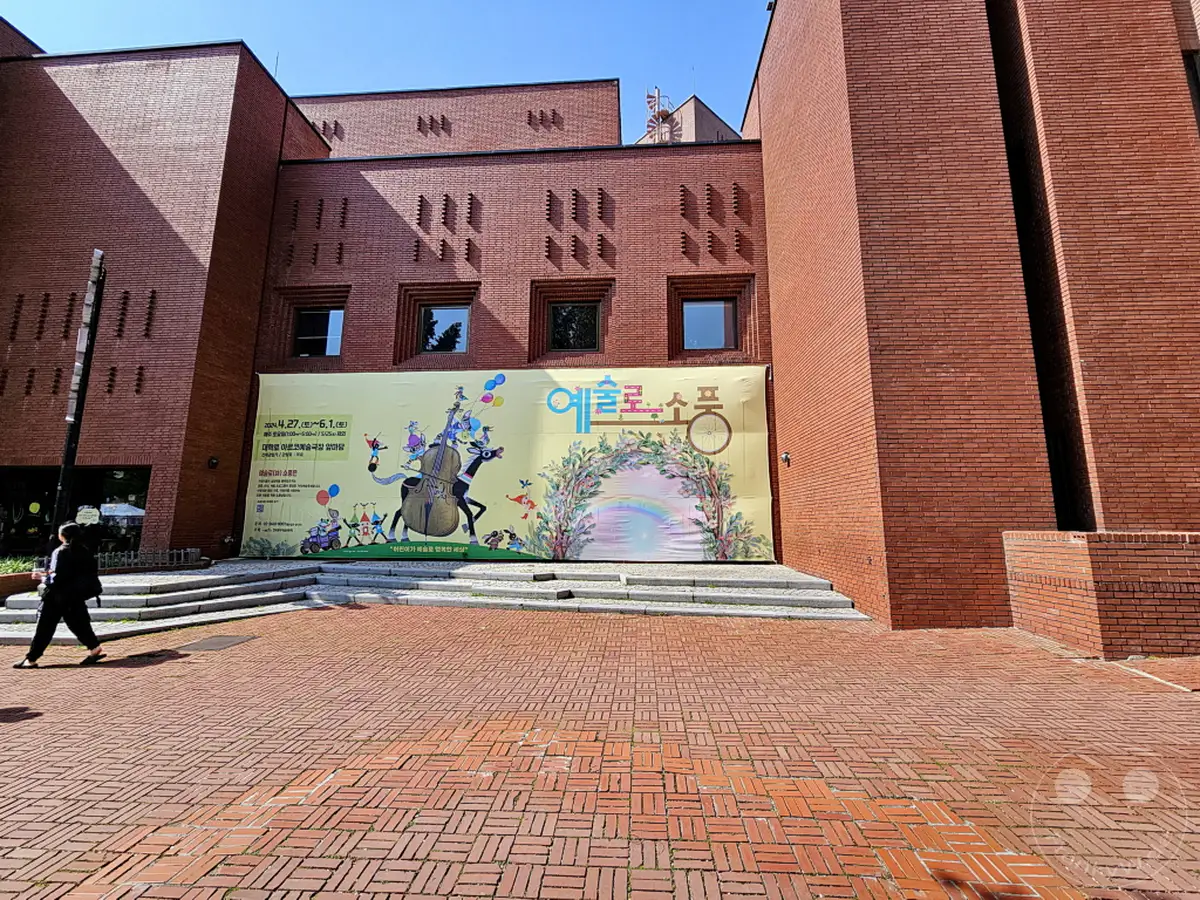Eine temporäre Ausstellungsbanner ist an der roten Backsteinfassade eines modernen Gebäudes in Südkorea angebracht. Die ungewöhnliche Architektur des Gebäudes im Daehangno district von Seoul steht im Kontrast zu den bunten Grafiken auf dem Banner. Eine Person geht auf dem gepflasterten Vorplatz an der Ausstellungsfläche vorbei.