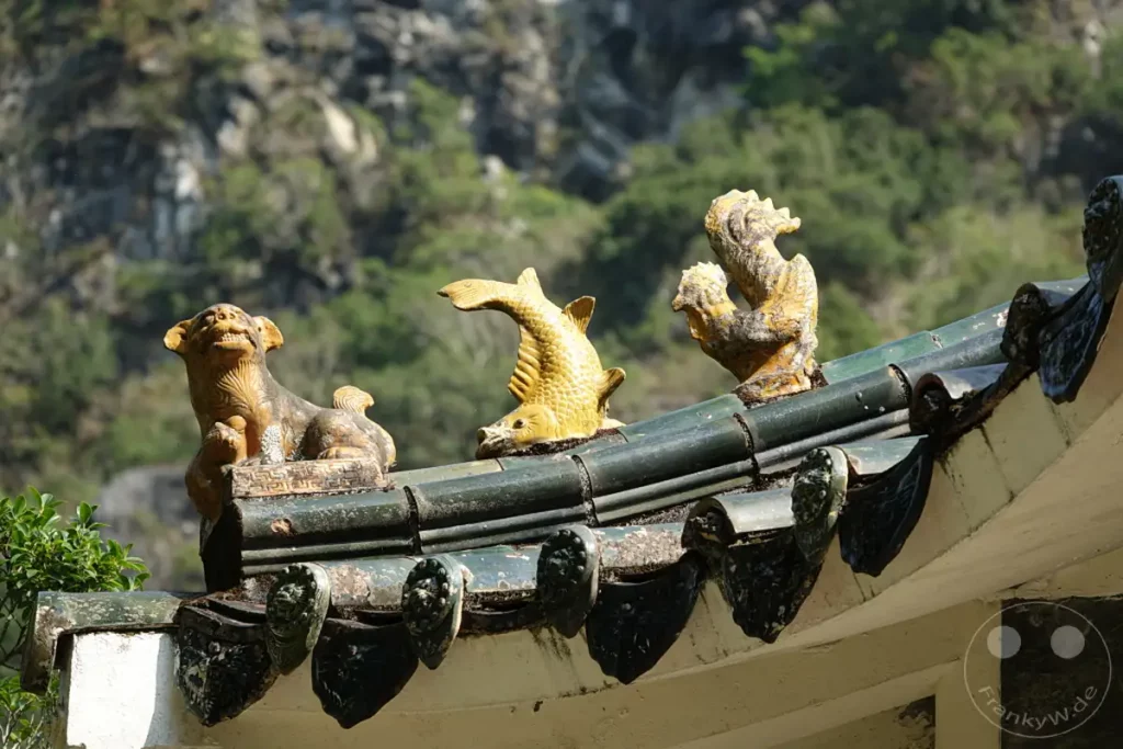 Figuren von einem Löwen, einem Fisch und einem Fabelwesen zieren die Dachziegel des Yuewangting Pavillons im Taroko Nationalpark in Taiwan. Die aufwendig gestalteten Skulpturen, teilweise verwittert und mit Moos überzogen, zeugen von traditioneller asiatischer Baukunst. Der sonnendurchflutete Hintergrund mit bewachsenen Berghängen vermittelt eine ruhige und fast mystische Atmosphäre.