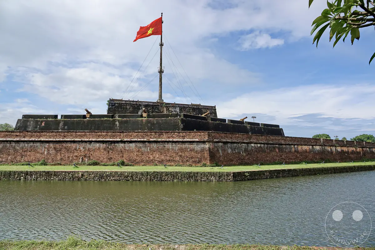 Eine wehende vietnamesische Flagge steht auf einem hohen Fahnenmast über der imposanten Zitadelle der Kaiserstadt Hue. Historische Kanonen sind auf den Zinnen platziert, die die mächtigen Backsteinmauern überblicken, während ein ruhter Wassergraben die Anlage umschließt. Der Himmel ist mit vereinzelten Wolken bedeckt, was der Szene eine majestätische und zugleich friedliche Atmosphäre verleiht.