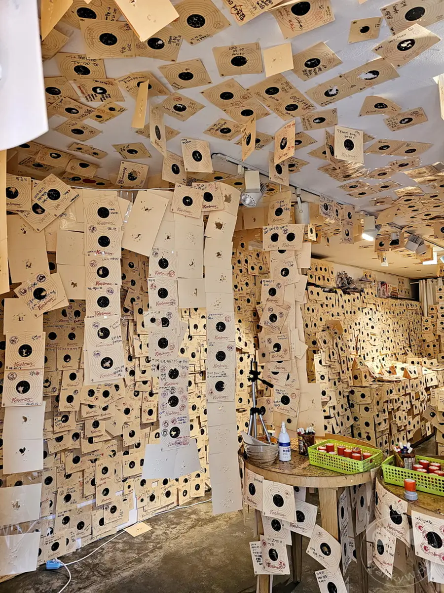 Eine Wand im Daehangno district in Seoul, Südkorea, ist vollständig mit Schießstand-Zielscheiben bedeckt, die unzählige Einschusslöcher aufweisen. Die Anordnung der Zielscheiben schafft eine dichte, fast überwältigende visuelle Collage aus persönlichen Erfolgen und dem visuellen Echo vergangener Schießübungen. Diese ungewöhnliche Präsentation verwandelt die Objekte einer Freizeitaktivität in eine bemerkenswerte und eindringliche Raumerfahrung.