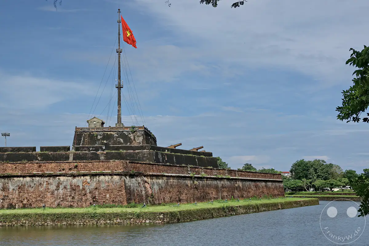 Die kaiserliche Zitadelle in Hue, Vietnam, erhebt sich imposant mit ihren alten Ziegelmauern und historischen Kanonen über dem umliegenden Wassergraben. Eine wehende vietnamesische Flagge auf dem hohen Fahnenmast symbolisiert die reiche Geschichte und kulturelle Bedeutung dieses Ortes. Die Szene strahlt eine ruhige und geschichtsträchtige Atmosphäre aus, eingefangen unter einem wolkenverhangenen blauen Himmel.