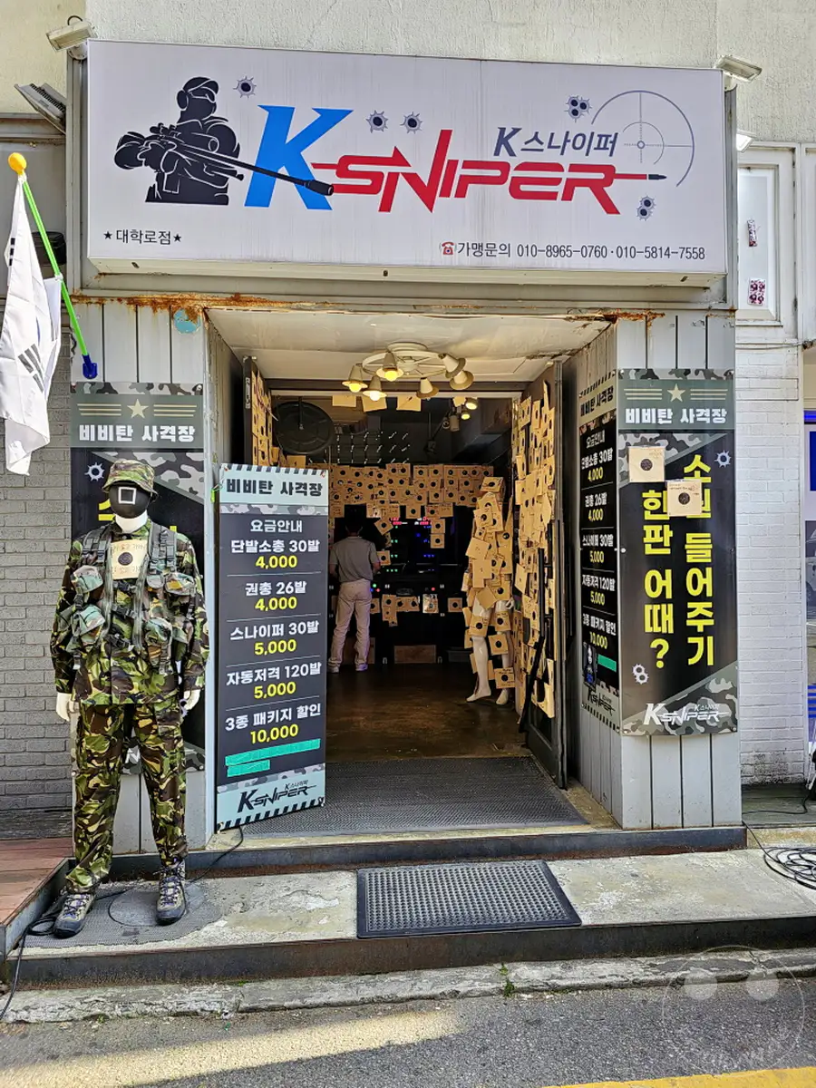 Der Eingang des K-Sniper Schießstands im Daehangno district von Seoul, Südkorea, ist mit Tarnkleidung und Werbetafeln geschmückt. Ein Mann in voller taktischer Ausrüstung steht als Schaufensterpuppe vor dem Laden, während drinnen die Schießanlage zu erkennen ist. Die Atmosphäre wirkt industriell und auf Action ausgelegt, typisch für eine Freizeiteinrichtung dieser Art.