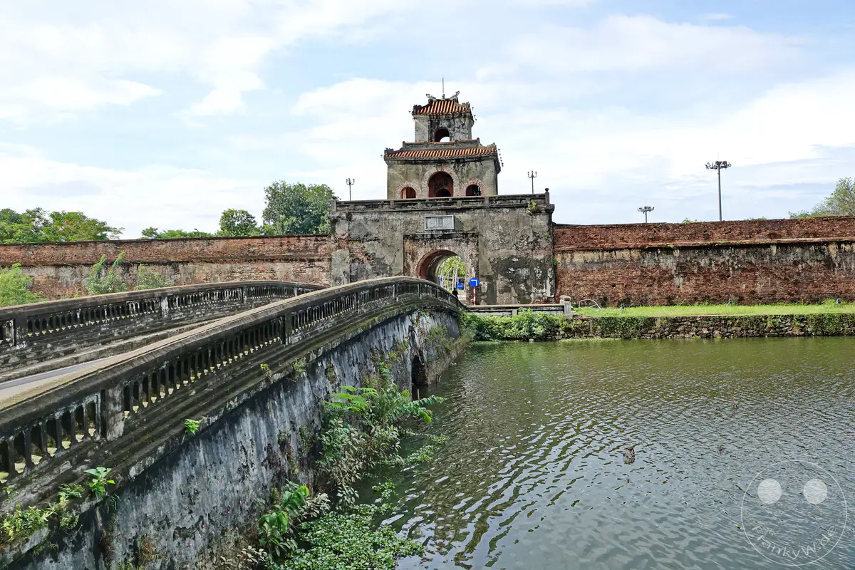 Die massive Backsteinmauer der Zitadelle von Hue, ein Meisterwerk der Annamitischen Architektur, umgibt die Kaiserstadt. Eine alte Steinbrücke mit kunstvollen Balustraden führt über den Wassergraben zur Festung. Die Szene strahlt eine ruhige, historische Atmosphäre aus, unterbrochen von vereinzelten Wolken am Himmel.