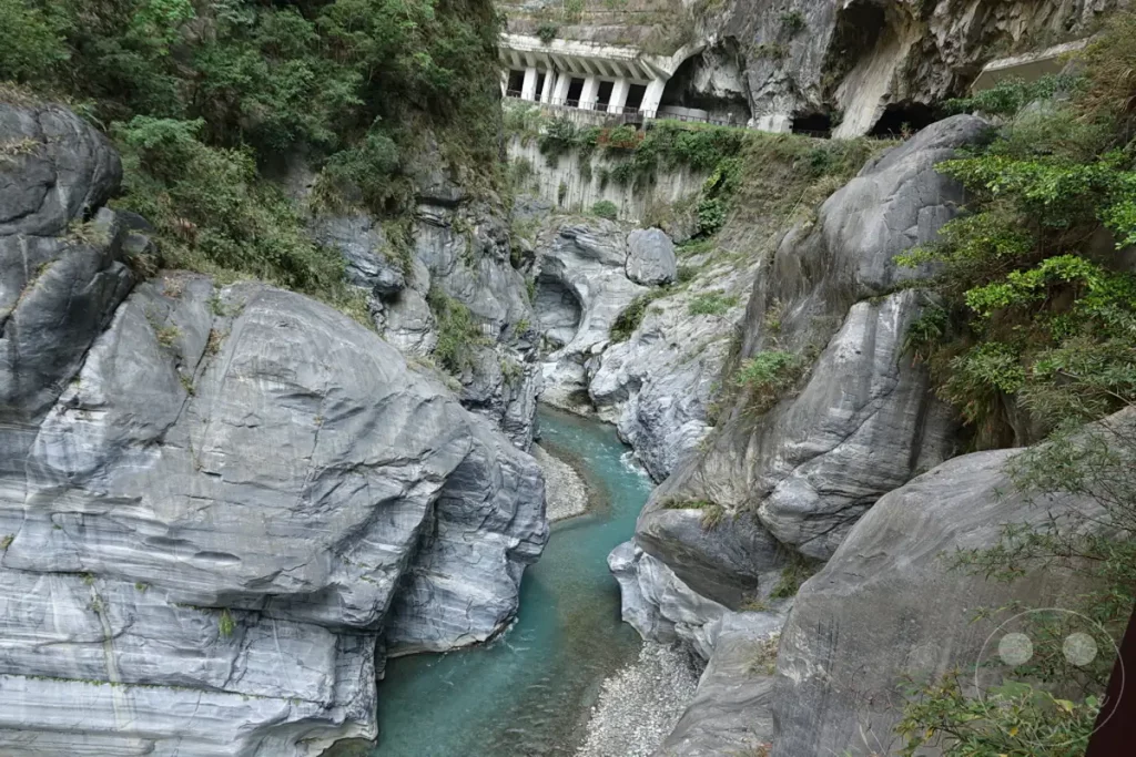 Eine türkisblaue Fluss schlängelt sich durch die tiefe, von steilen Felswänden gesäumte Taroko-Schlucht im Taroko Nationalpark in Taiwan. Die gewaltigen Marmorformationen zeugen von der erodierenden Kraft des Wassers über Jahrtausende, während im Hintergrund eine künstlich angelegte Struktur, wahrscheinlich ein Teil des Tunnel of Nine Turns, in die Bergwand gebaut ist. Die Szenerie strahlt eine majestätische und zugleich raue Schönheit aus, die von der beeindruckenden Naturkraft und menschlichen Ingenieurskunst geprägt ist.
