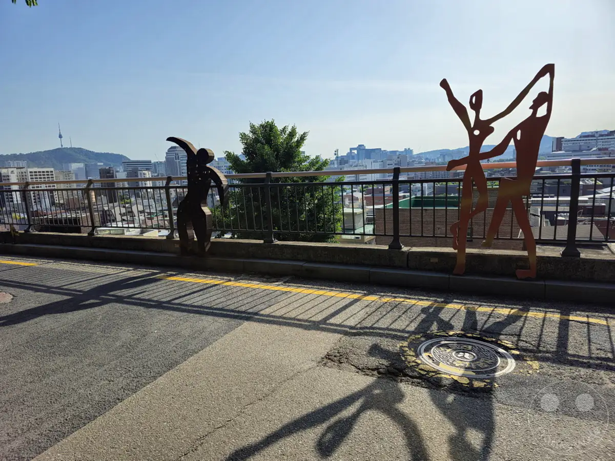 Silhouette-Skulpturen von tanzenden Figuren säumen einen Geländerweg mit Blick auf die Stadtlandschaft von Seoul. Im Hintergrund erheben sich Gebäude und ein Fernsehturm vor einem klaren blauen Himmel. Die Szene vermittelt ein Gefühl von Kunst im öffentlichen Raum und urbaner Erkundung im berühmten Ihwa Mural Village.