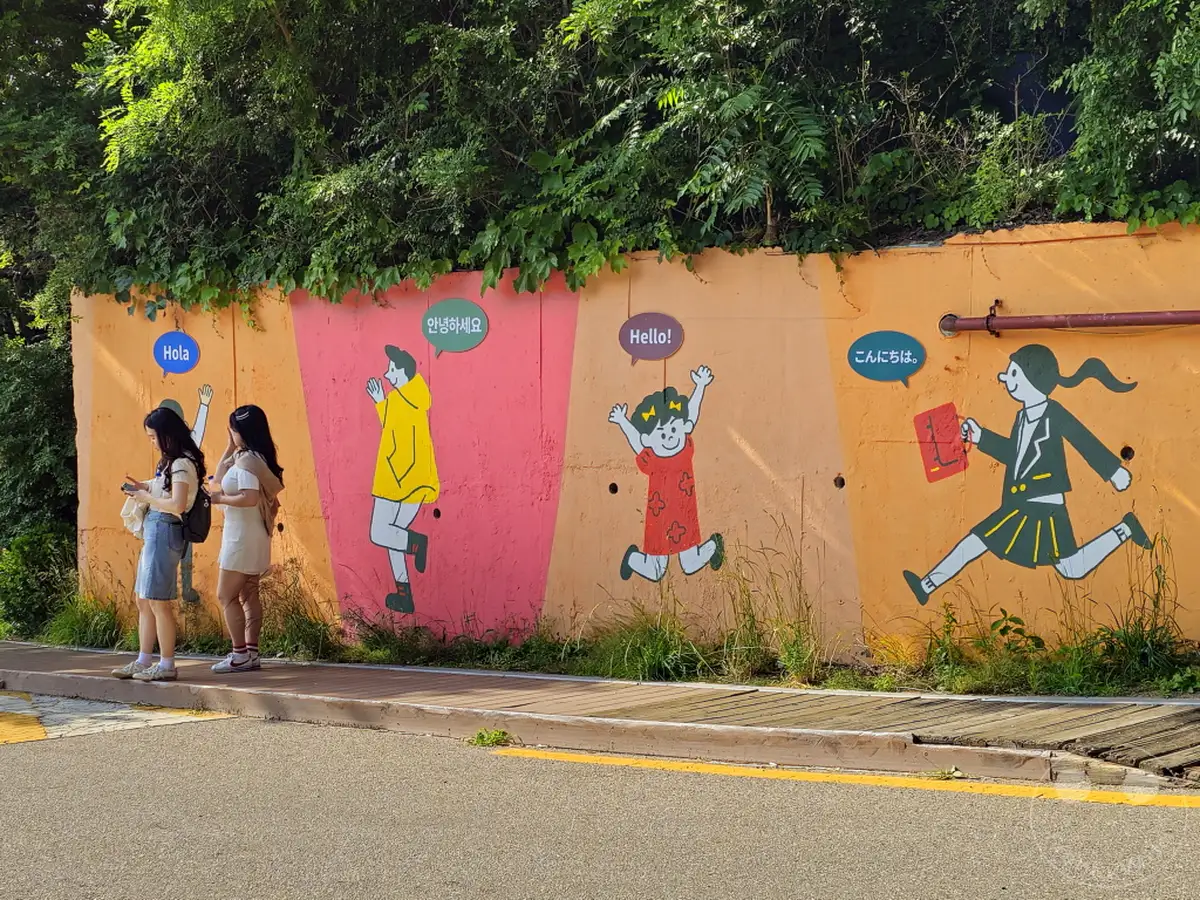 Die bunte Wandmalerei in Südkoreas Ihwa Mural Village in Seoul begrüßt Besucher in verschiedenen Sprachen. Kindliche Darstellungen mit Grußformeln wie 'Hola', '안녕하세요' und 'Hello!' schaffen eine fröhliche und einladende Atmosphäre. Zwei junge Frauen stehen vor der Wand und betrachten die Kunstwerke.