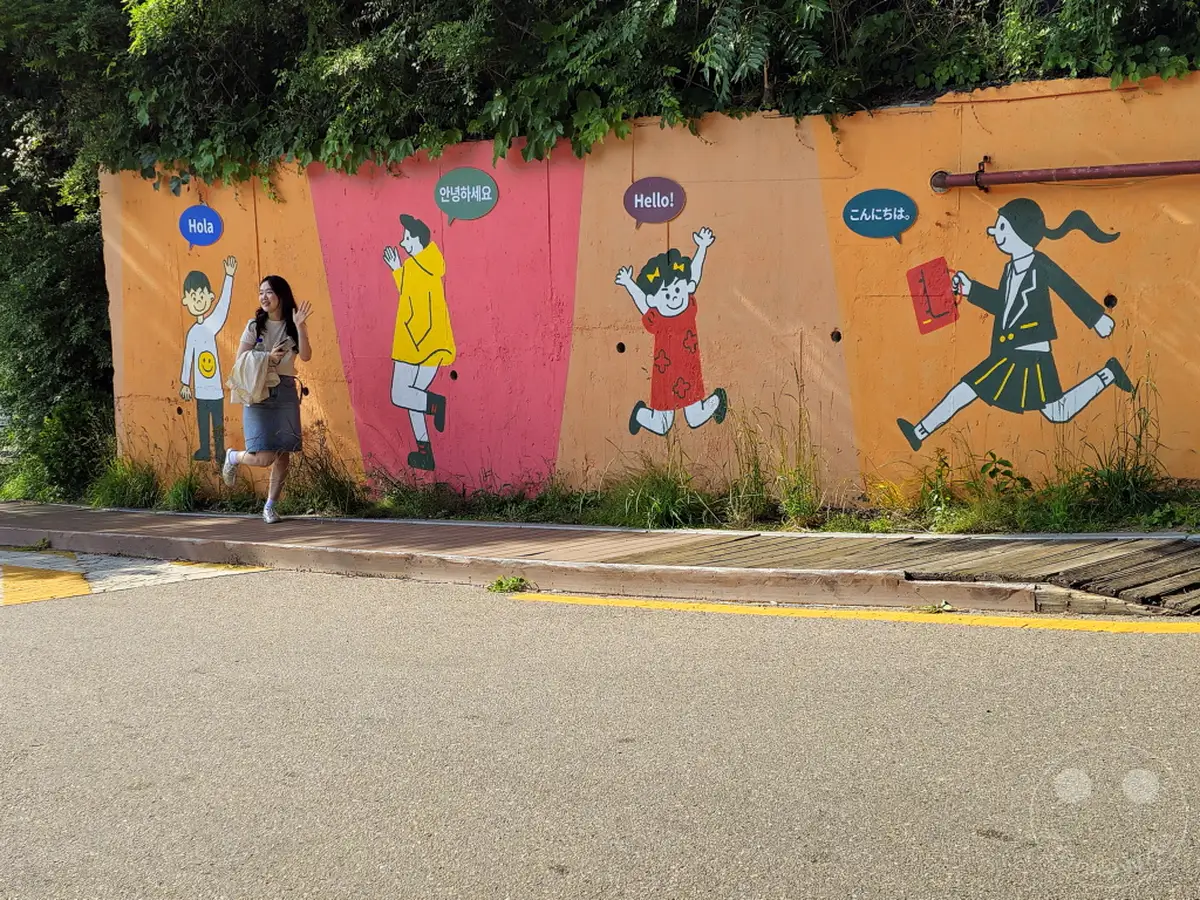 Eine junge Frau steht lächelnd vor einer bunten Wandmalerei im Ihwa Mural Village in Seoul, Südkorea. Die Wand ist mit verschiedenen Figuren und Grußformeln in unterschiedlichen Sprachen verziert, was eine fröhliche und internationale Atmosphäre schafft. Die Szene vermittelt ein Gefühl von Entdeckung und kultureller Vielfalt in einer urbanen Umgebung.