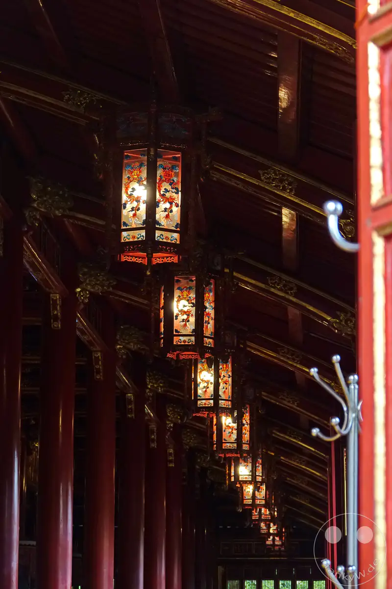 Eine Reihe kunstvoll verzierter Lampions mit Drachenmotiven beleuchtet einen langen, roten Korridor innerhalb der Kaiserstadt in Hue, Vietnam. Die traditionelle Architektur, geprägt von roten Säulen und goldenen Verzierungen, schafft eine feierliche und historische Atmosphäre. Das warme Licht der Lampions fällt auf die polierten Oberflächen und betont die Detailverliebtheit der königlichen Anlage.