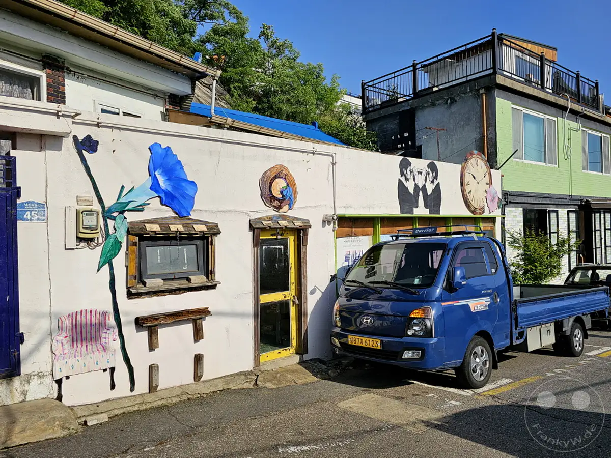 In Südkoreas Hauptstadt Seoul, genauer gesagt im Ihwa Mural Village, belebt eine großflächige Wandmalerei einer blauen Blume die Fassade eines Hauses. Ein blauer Lieferwagen steht geparkt davor und vervollständigt das bunte Straßenbild. Die Szene strahlt eine lebendige und künstlerische Atmosphäre aus, die typisch für dieses Viertel ist.