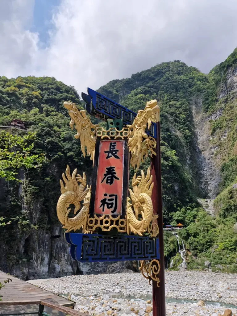 Die chinesischen Schriftzeichen "長春祠" (Changchun Shrine) sind auf einem roten Schild über einer steinigen Flusslandschaft in Taiwan angebracht. Majestätische grüne Berge und Felswände umgeben den Schrein, während ein Wasserfall in der Ferne in die Tiefe stürzt. Die Szenerie strahlt eine friedliche und zugleich beeindruckende Atmosphäre der Natur aus.