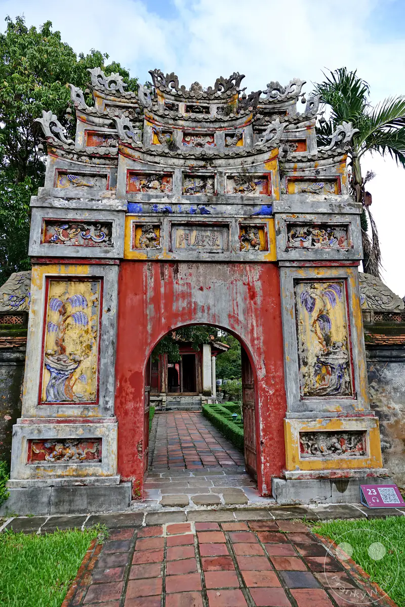 Ein prächtiges Tor zur Kaiserstadt Hue in Vietnam, das mit aufwendigen Verzierungen und verwitterten Farben glänzt. Die Architektur zeugt von reicher Geschichte und traditionellem Handwerk, während der umliegende Garten und der gepflasterte Weg zur Erkundung einladen. Die Szene strahlt eine Atmosphäre vergangener Pracht und stiller Erhabenheit aus.
