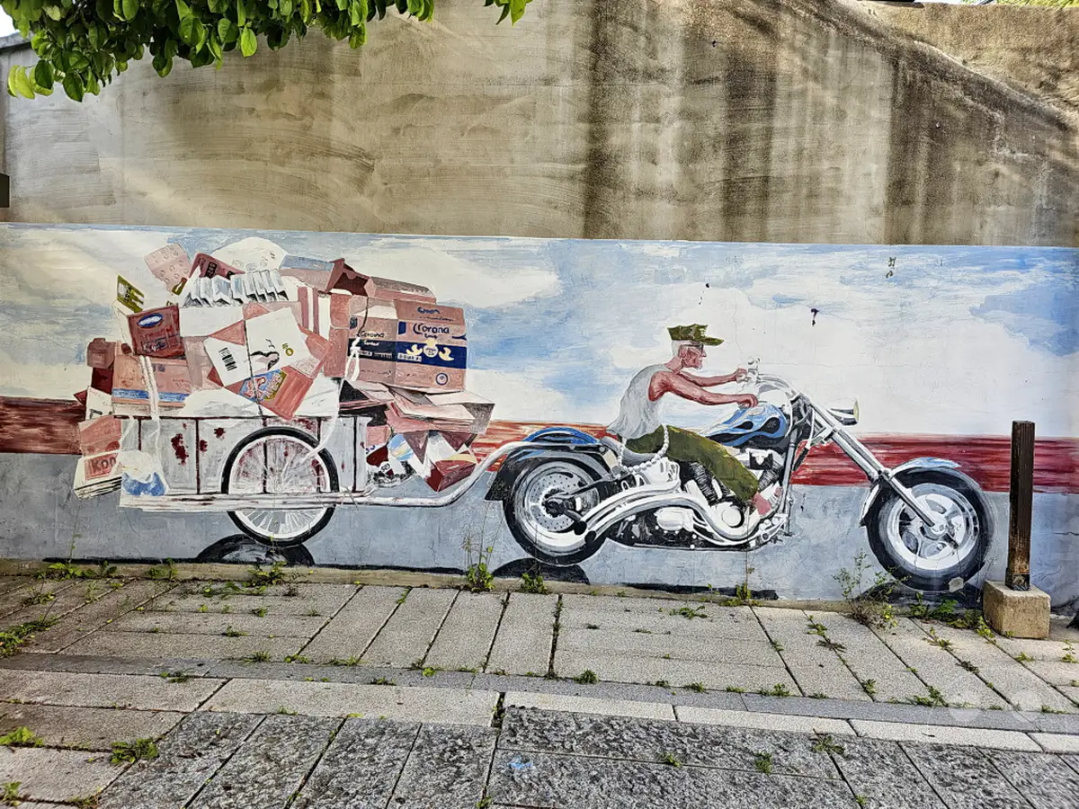 Eine detailreiche Wandmalerei in Seoul im Ihwa Mural Village zeigt einen Mann auf einem Motorrad, der einen mit vielen Kisten beladenen Anhänger zieht. Die bunte und lebendige Darstellung vermittelt eine geschäftige Atmosphäre, die typisch für die künstlerische Gestaltung des Viertels ist.