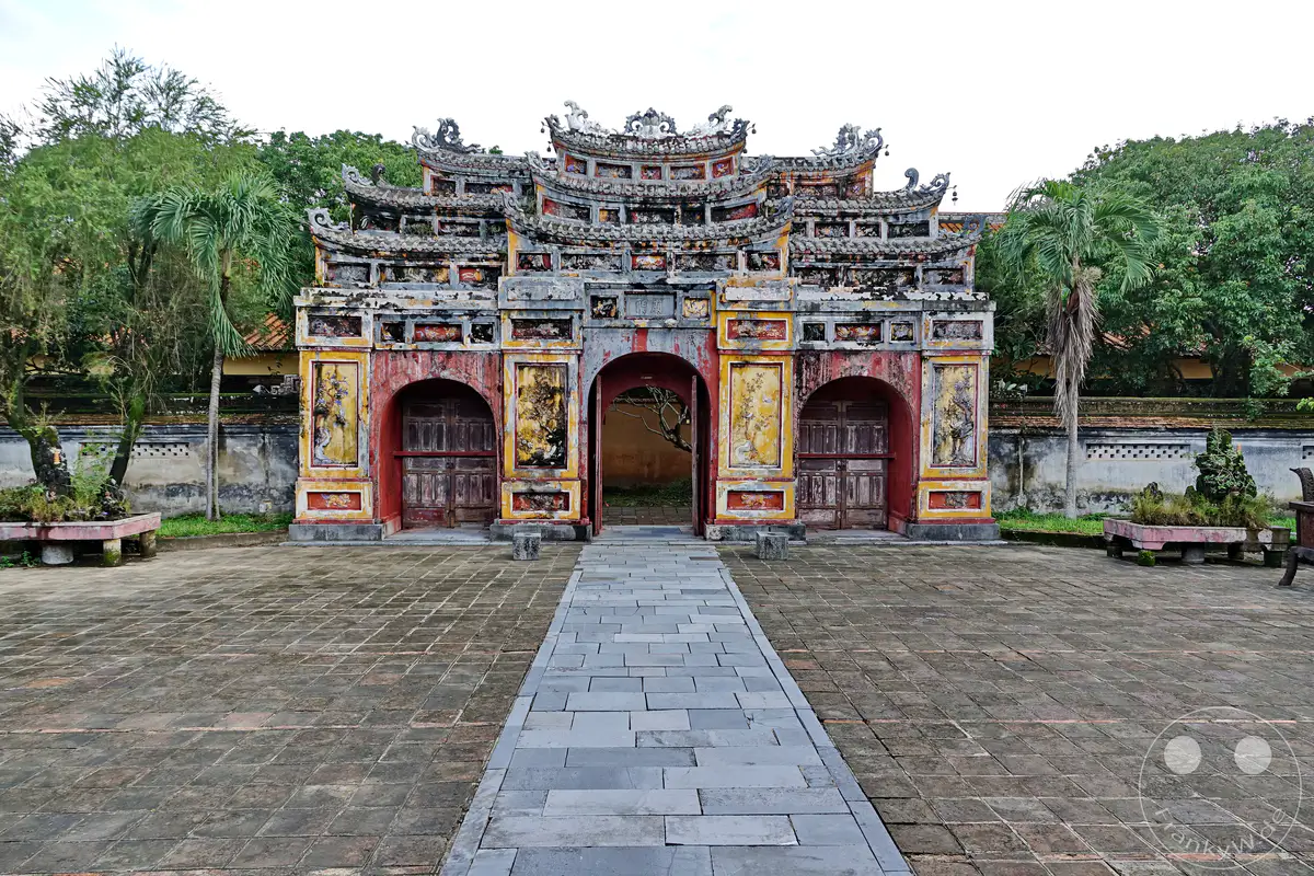Ein imposantes Tor, Teil der Kaiserlichen Stadt in Hue, Vietnam, mit detaillierter Architektur und Verzierungen. Die rote Fassade und die alten Holztüren verleihen dem Bauwerk Geschichte und Charakter. Ein gepflasterter Weg führt durch den Innenhof, umgeben von üppiger Vegetation und alten Mauern.
