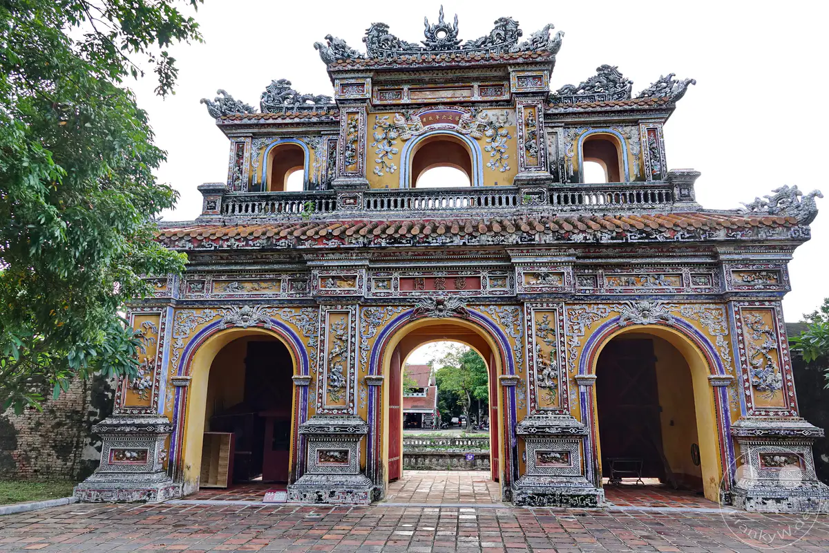 Ein imposantes, detailreich verziertes Tor der Zitadelle von Hue in Vietnam. Die ornate Fassade, geschmückt mit Drachenfiguren und Mustern, führt zu einer Anlage, die auf eine reiche kaiserliche Vergangenheit verweist. Die Szene strahlt eine Atmosphäre historischer Erhabenheit und kultureller Tiefe aus.