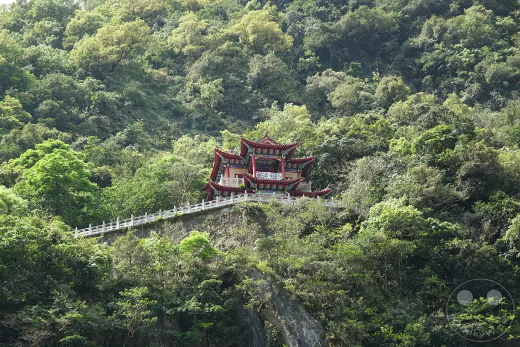 Der Changchun-Schrein in Taiwan, eingebettet in die üppige Vegetation des Taroko-Nationalparks, thront auf einer Klippe über einem bewaldeten Tal. Die traditionelle asiatische Architektur des Schreins mit seinen roten Dächern bildet einen markanten Kontrast zur satten grünen Bergkulisse. Die Szene strahlt eine friedliche und erhabene Atmosphäre aus, die die Harmonie zwischen Kultur und Natur betont.