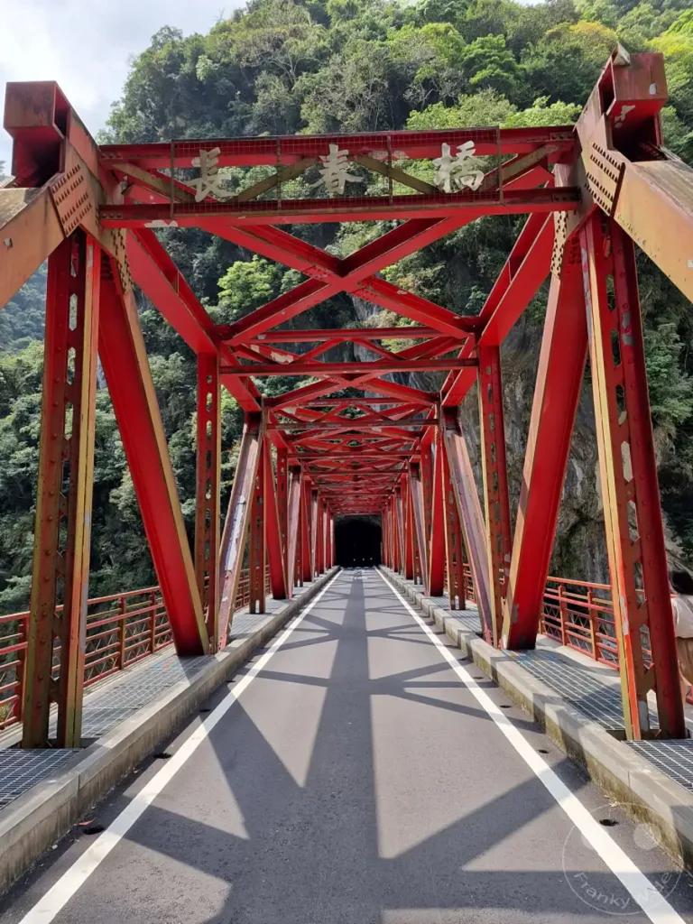 Die ikonische rote Changchun-Brücke führt durch den Taroko Nationalpark in Taiwan. Die Stahlkonstruktion wirft markante Schatten auf die asphaltierte Straße, die in einen dunklen Tunnel mündet. Die üppige grüne Berglandschaft bildet einen dramatischen Kontrast zur künstlichen Struktur der Brücke.