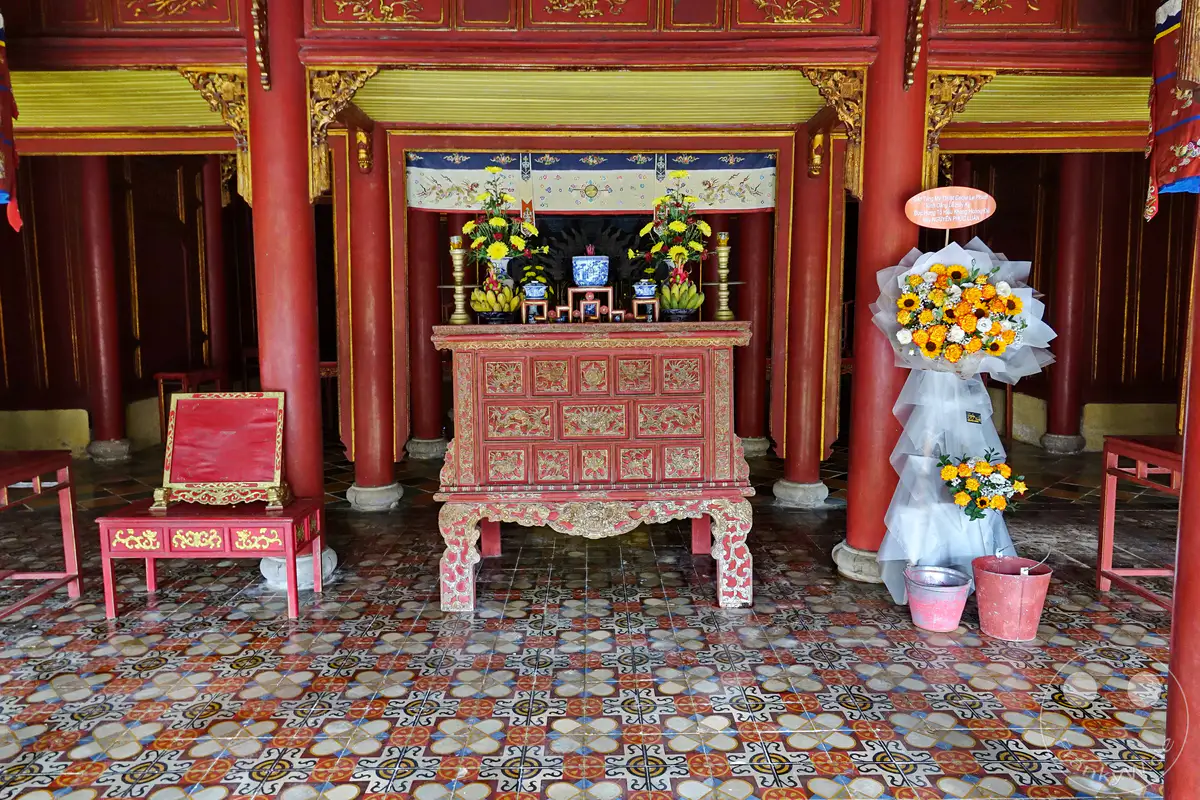In der kaiserlichen Zitadelle von Hue, Vietnam, steht ein reich verzierter Altar im Herzen eines traditionellen Gebäudes. Die aufwendigen roten und goldenen Verzierungen sowie die symmetrisch arrangierten Blumen und Früchte vermitteln eine Atmosphäre von Ehrfurcht und kultureller Bedeutung. Die detailreichen Bodenfliesen und die mächtigen roten Säulen verstärken den Eindruck von historischer Pracht und spiritueller Stille.