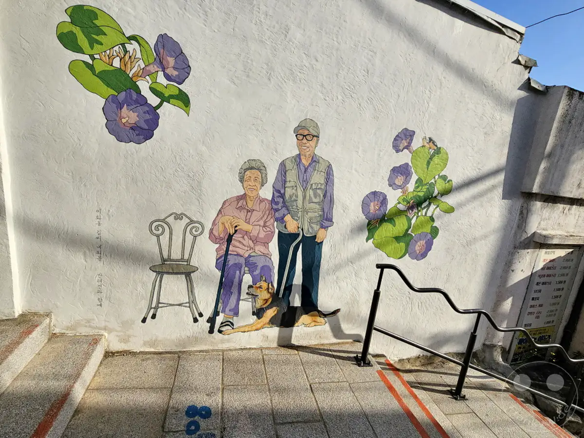 Eine farbenfrohe Wandmalerei im Ihwa Mural Village in Seoul zeigt ein älteres Ehepaar mit ihrem Hund, umgeben von blauen Morning Glory Blumen und einem stilisierten Stuhl. Die Szene strahlt eine warme und nostalgische Atmosphäre aus, die das ländliche Leben und die Gemeinschaft hervorhebt. Das Kunstwerk ist auf einer rauen weißen Wand angebracht, die den Charme des Viertels unterstreicht.