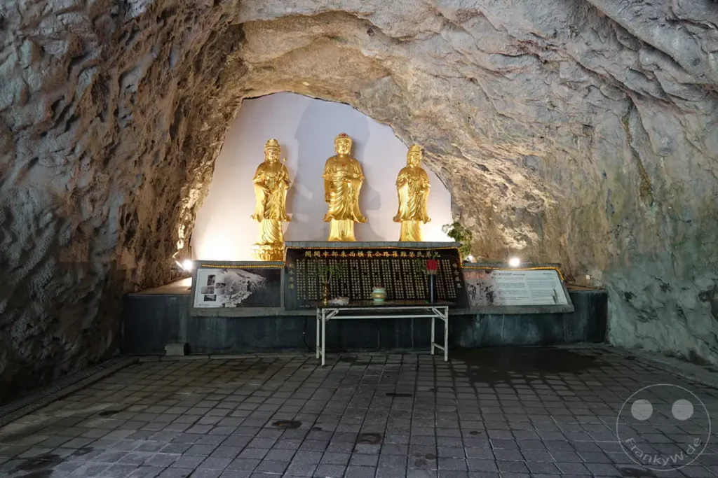 Im Taroko Nationalpark in Taiwan befindet sich der Changchun-Schrein, der tief in einer Höhle liegt. Goldene Buddha-Statuen stehen auf einem Podest vor einer Gedenktafel mit chinesischen Schriftzeichen, beleuchtet von Scheinwerfern, die die raue Höhlenarchitektur betonen. Die Atmosphäre ist ruhig und andächtig, inmitten der natürlichen Formationen des Nationalparks.