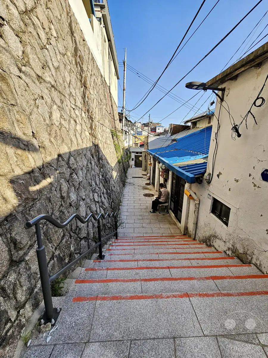 Eine steile, mit roten Markierungen versehene Steintreppe führt durch eine schmale Gasse in Südkorea, Seoul. Links ragt eine grob behauene Steinmauer auf, rechts säumen weiße Hauswände mit blauen Dächern und einem Mann auf einer Bank den Weg. Der strahlend blaue Himmel überspannt die Szene und verleiht ihr eine friedliche und alltägliche Atmosphäre.