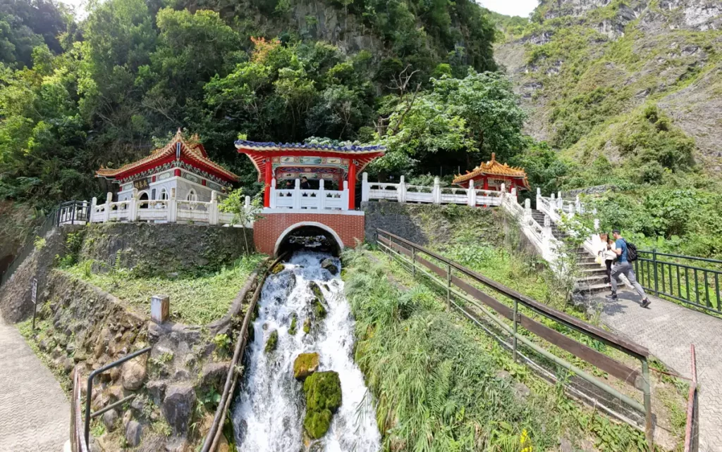 Ein malerischer Wasserfall stürzt von den Bergen herab und fließt unter einem traditionellen chinesischen Tempel hindurch. Grüne Vegetation und steile Berghänge umgeben die Anlage, die eine friedliche Atmosphäre ausstrahlt. Zwei Besucher erkunden den Weg zu den Tempeln und über eine Treppe, die sich durch die Landschaft schlängelt.
