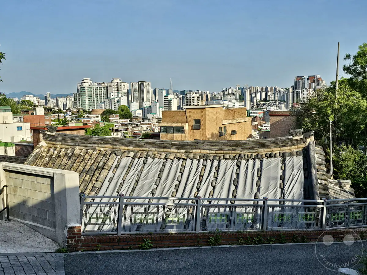 Ein Panoramablick über das Ihwa Mural Village in Seoul, Südkorea, zeigt einen faszinierenden Kontrast zwischen traditionellen koreanischen Dächern und der modernen Stadtlandschaft im Hintergrund. Die detaillierte Textur der alten Ziegel und die schützenden Planen über den Dächern stehen im Dialog mit den hoch aufragenden Gebäuden der Metropole. Die Szene vermittelt eine ruhige, kontemplative Atmosphäre, die zum Verweilen und Betrachten einlädt.