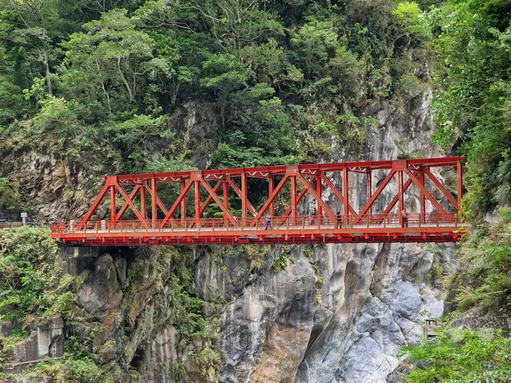 Die ikonische rote Changchun-Brücke überspannt eine tiefe Schlucht im Taroko Nationalpark in Taiwan. Steile, bewachsene Felswände und üppige Vegetation umrahmen das imposante Bauwerk und schaffen eine dramatische Kulisse. Die Szene strahlt eine friedliche, aber zugleich überwältigende Schönheit der Natur aus.