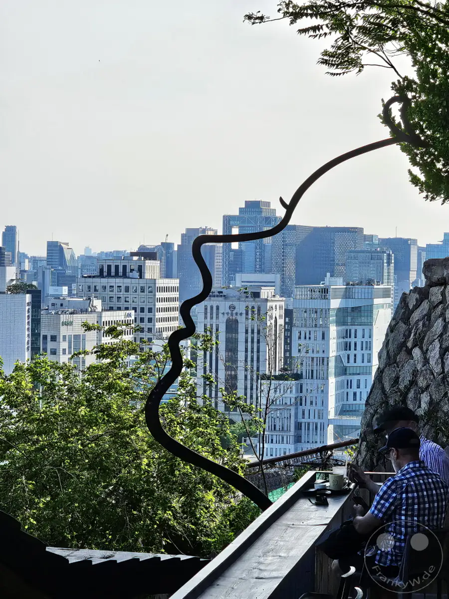 Ein geschwungenes, schlangenartiges Kunstwerk ragt von oben ins Bild und formt schemenhaft eine menschliche Profilansicht. Im Hintergrund erstreckt sich die moderne Stadtkulisse von Seoul, Südkorea, überragt von zahlreichen Hochhäusern. Auf einer Holzterrasse eines Cafés im Ihwa Mural Village sitzen zwei Personen und genießen die Aussicht, was eine entspannte Atmosphäre schafft.