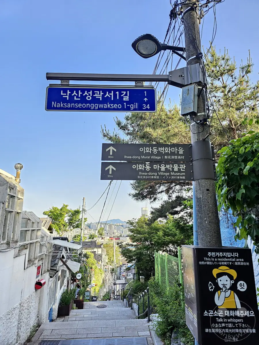 Eine steile Treppe führt durch das Ihwa Mural Village in Seoul, Südkorea, gesäumt von charmanten Häusern und Kunstwerken. Wegweiser und Hinweisschilder informieren über Sehenswürdigkeiten und Verhaltensregeln in der malerischen Gasse. Der Blick reicht über die Stadt und vermittelt eine ruhige, einladende Atmosphäre.
