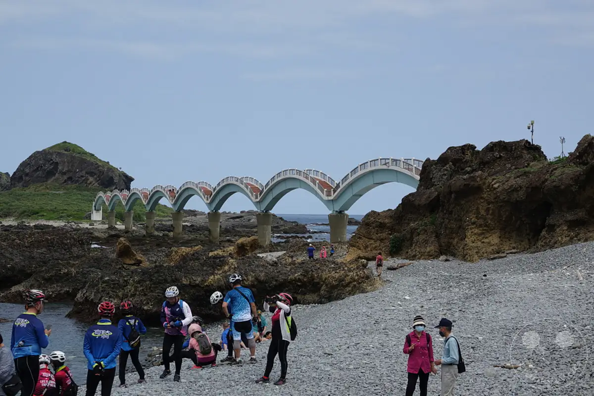 Auf einer kiesbedeckten Küstenlinie in Taiwan stehen Radfahrer und Touristen vor der markanten Sanxiantai Arch Bridge, einer mehrbogigen Brücke, die über das Meer führt. Die felsige Landschaft und das weite Meer bilden eine dramatische Kulisse für die Besucher, die die Aussicht genießen. Die Stimmung ist entspannt und touristisch, mit Menschen, die die einzigartige Architektur und die natürliche Schönheit der Umgebung erkunden.