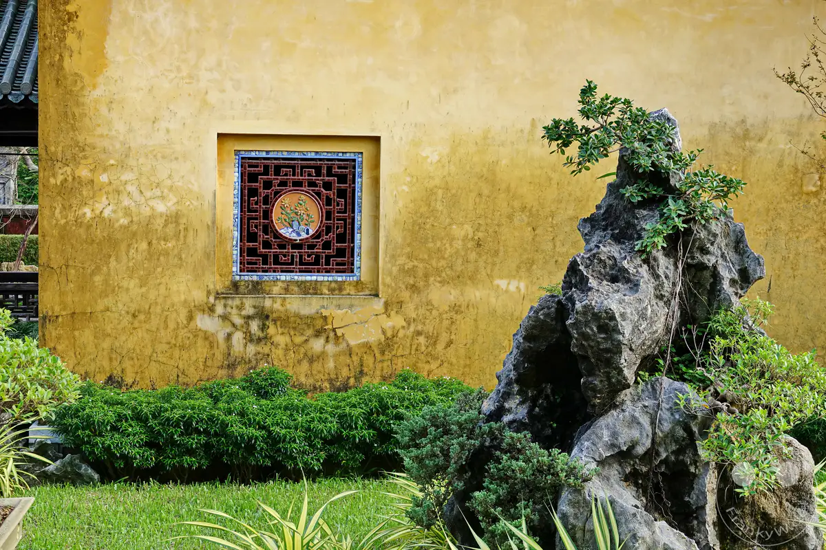 Die gelbe Mauer der Zitadelle in Hue, Vietnam, mit einem kunstvoll verzierten traditionellen Fenster, das von einem üppigen Felsengarten mit immergrünen Pflanzen umgeben ist. Die Szene strahlt eine Atmosphäre von Ruhe und historischer Schönheit aus, die die architektonische Eleganz und die natürliche Landschaft vereint. Dieser Anblick bietet einen Einblick in die reiche Kultur und das Erbe des Kaiserpalastes.