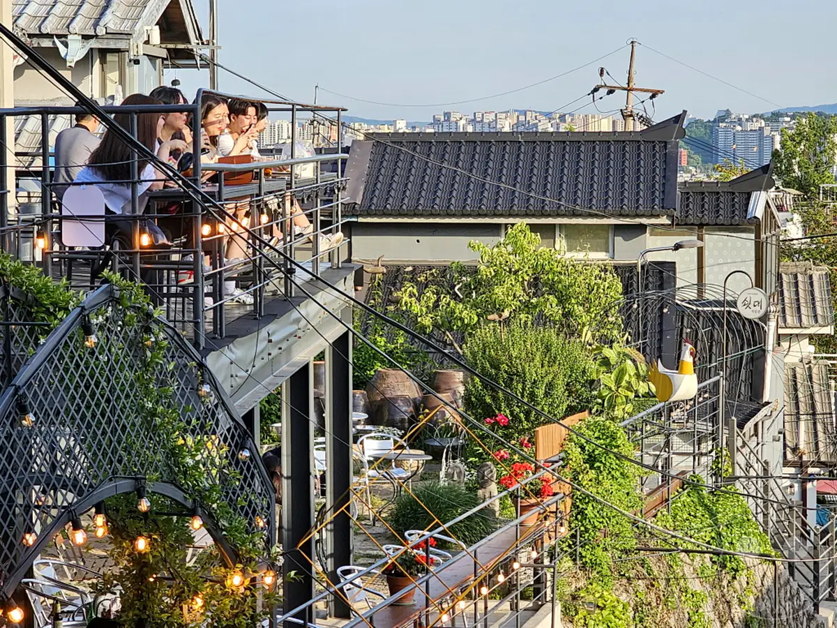 Eine Gruppe von Freunden genießt den Abend in einem Rooftop-Café in Seoul, Südkorea, mit Blick auf das belebte Stadtbild. Die stimmungsvolle Beleuchtung und die üppige Begrünung schaffen eine entspannte Atmosphäre. Dieses Bild fängt die lebendige Kultur und die charmanten Ausblicke des Ihwa Mural Village ein.