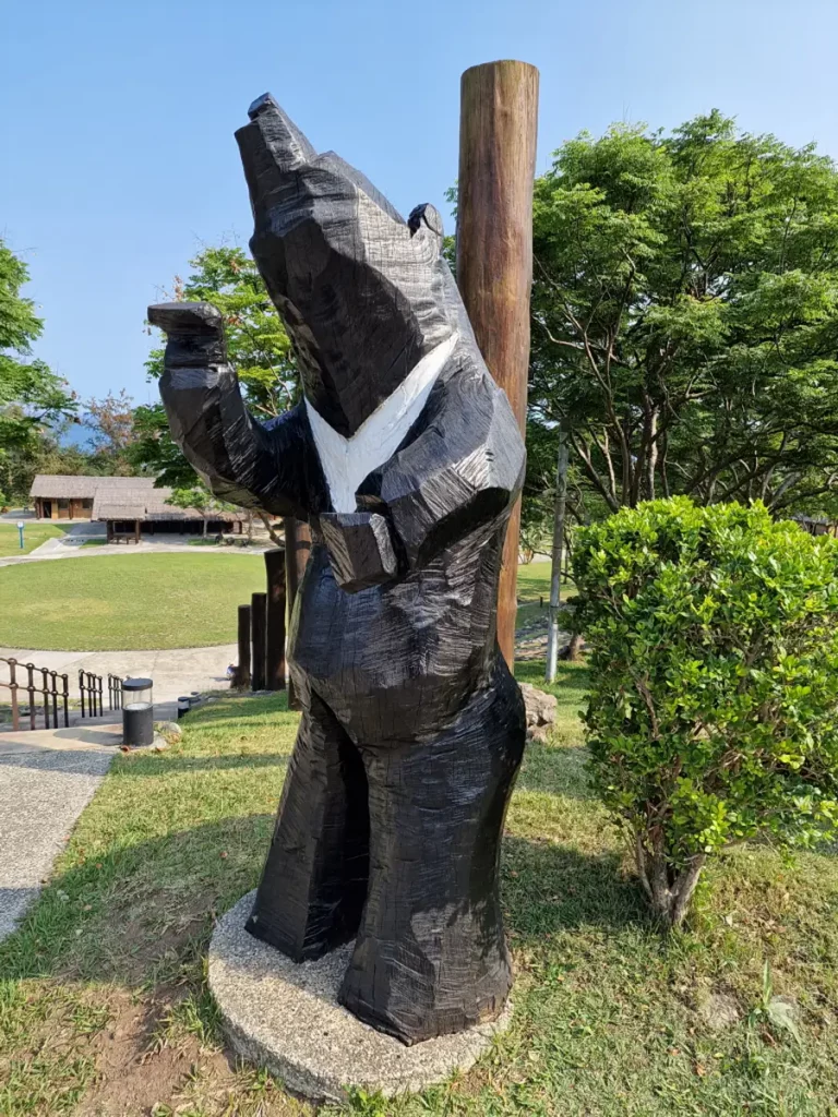 Eine geschnitzte Bärenskulptur in Anzug steht aufrecht in der Nähe eines Baumstammes. Die Szene spielt im Amis Folk Center in Taiwan, wo traditionelle Architektur und Kunstwerke inmitten einer gepflegten Grünanlage zu sehen sind. Die sonnige Atmosphäre und die ländliche Umgebung laden zum Erkunden ein.