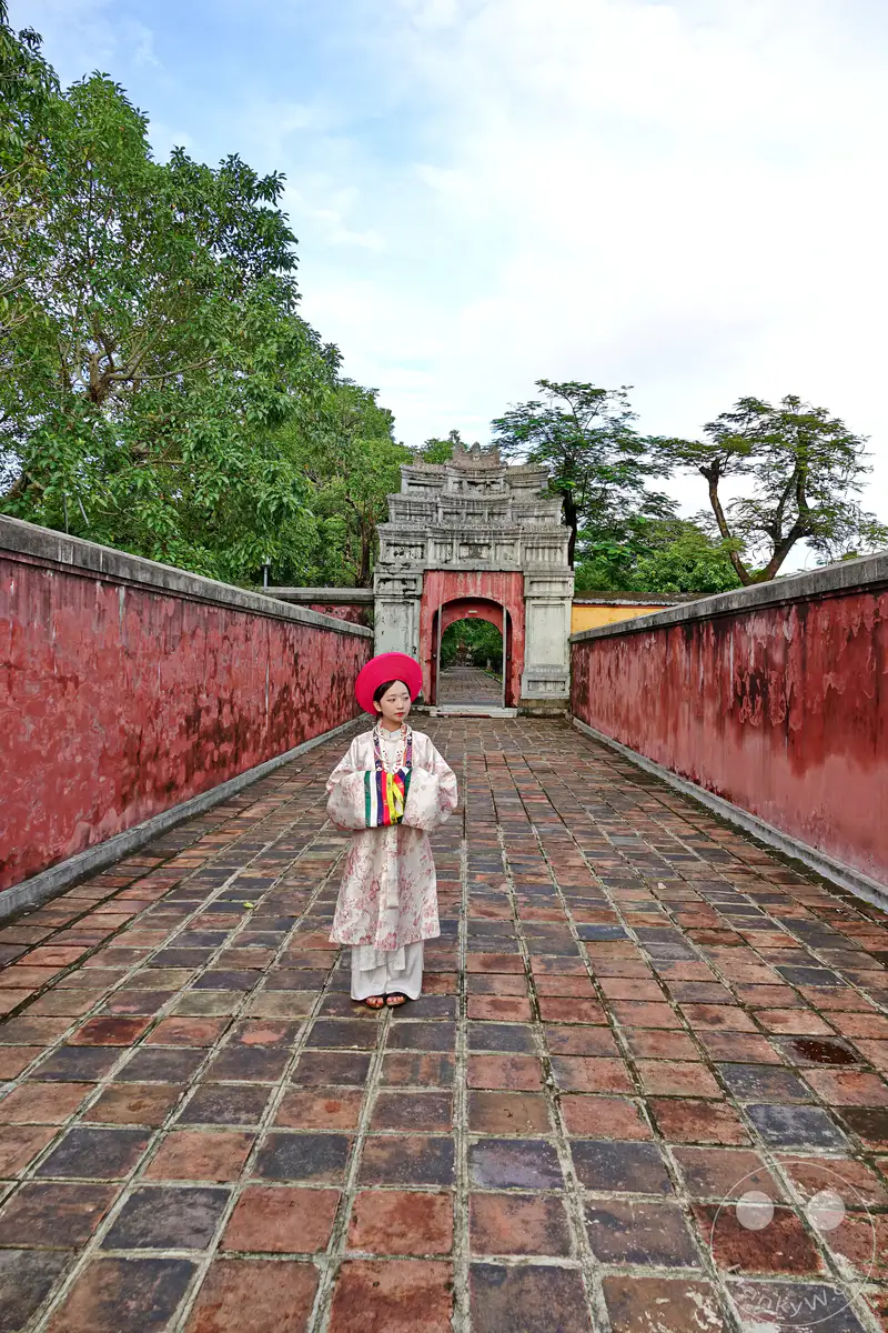 Eine junge Frau in traditioneller Kleidung steht auf einem mit Ziegelsteinen gepflasterten Weg vor einem imposanten Torbogen in der Kaiserstadt Hue, Vietnam. Die roten Mauern und die Architektur des Bauwerks vermitteln eine historische und feierliche Atmosphäre. Das Bild fängt einen Moment der Ruhe und kulturellen Verbindung in dieser historischen Stätte ein.